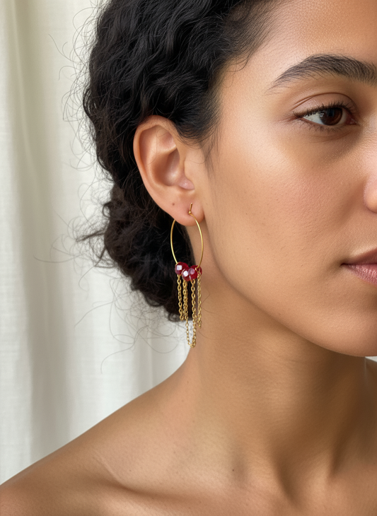 Close-up of a woman wearing gold hoop earrings with red gemstones against a neutral background